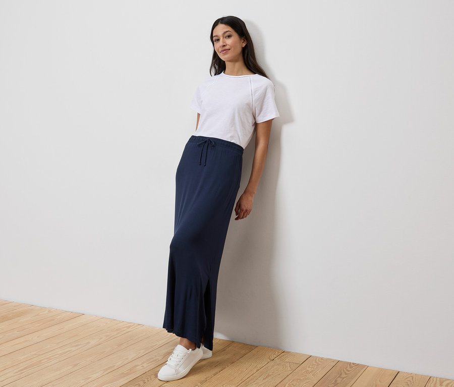 Nő támaszkodik a falnak, fehér pólót, hosszú, sötétkék szoknyát és fehér tornacipőt visel.