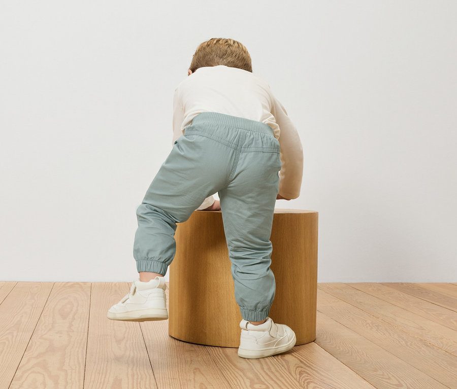 Egy gyerek felmászik egy fa zsámolyra, hosszú ujjú felsőt és világoskék nadrágot visel.