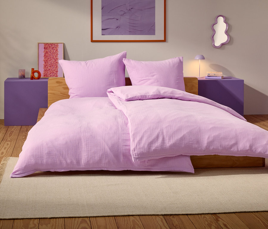 Egy lila ágy két párnával és egy takaróval egy bézs szőnyegen áll egy hálószobában.