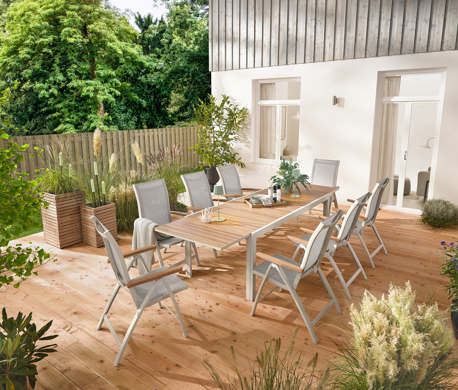 Egy kb. 200–300 cm hosszú, kihúzható alumínium kerti asztal »Liska« áll egy fa teraszon, amelyet hat darab, állítható háttámlás »Liska« magas támlás szék vesz körül. Két darab »Tinus« ültetőláda és 5 darab LED-es kerti fáklya is látható.