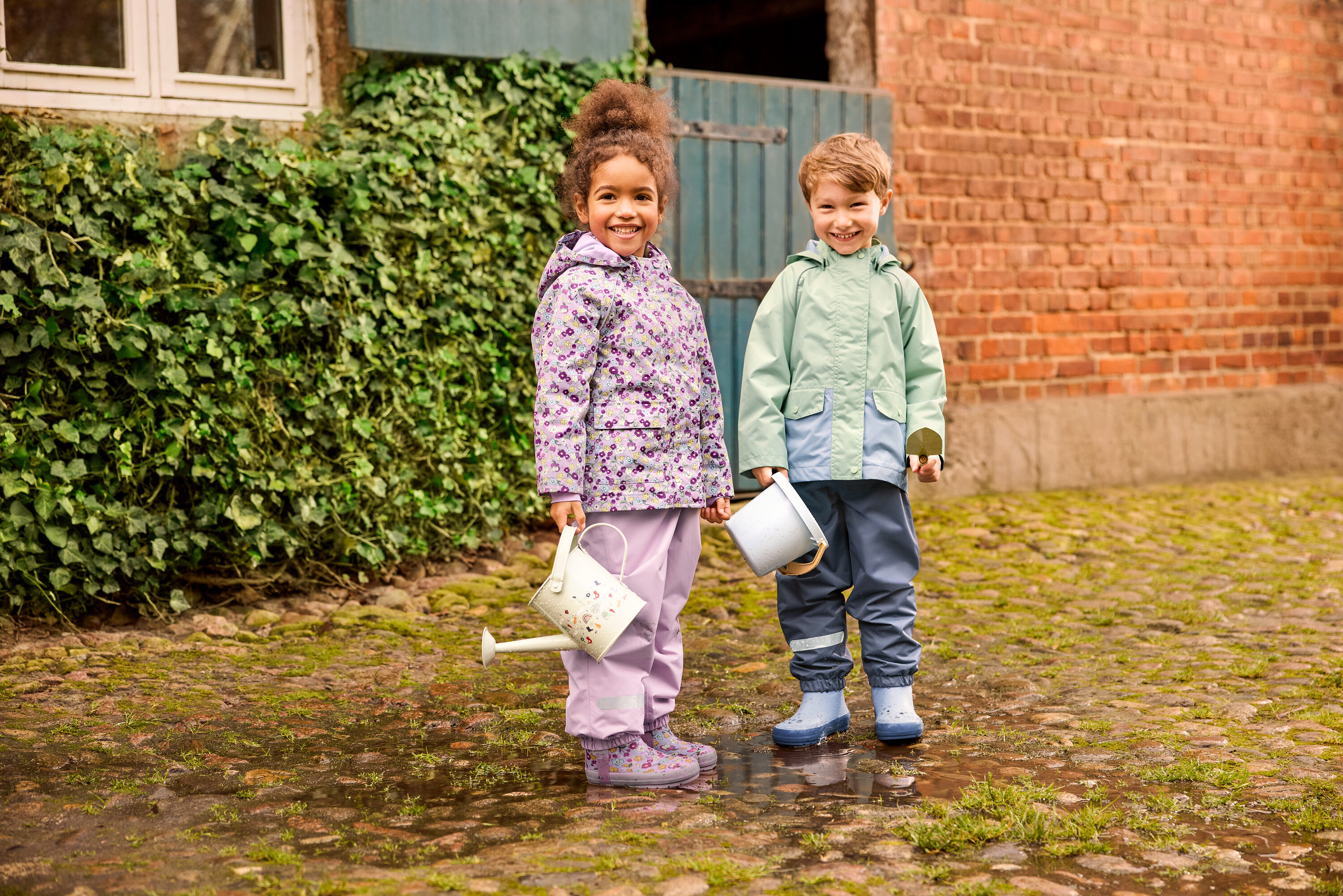 Két gyerek áll kint: egy virágos gyerek esőkabátot és virágos gyerek gumicsizmát viselő lány, és egy zöld gyerek esőkabátot és kék gyerek gumicsizmát viselő fiú. Mindketten gyerek esőnadrágot viselnek.