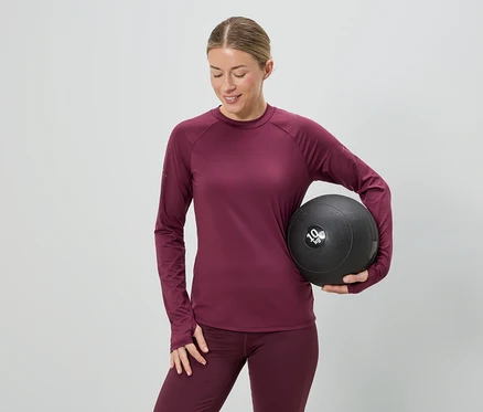 Bordó sportruházatot viselő nő egy 10 kg-os medicinlabdát tart szürke háttér előtt.