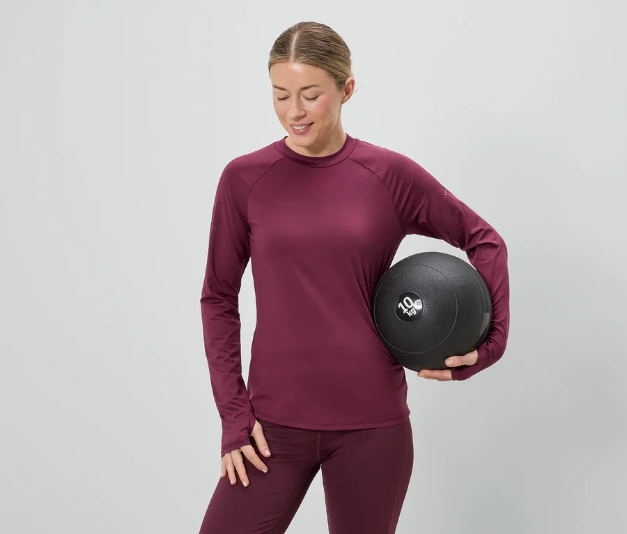 Bordó sportruházatot viselő nő egy 10 kg-os medicinlabdát tart szürke háttér előtt.