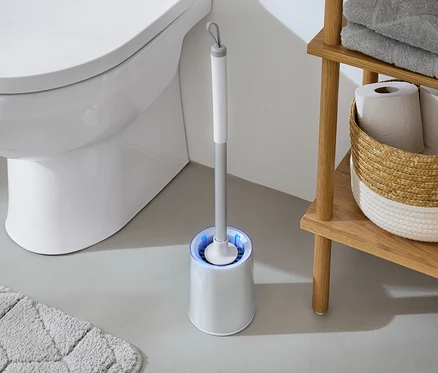 A fürdőszobában egy fehér WC áll egy kék fényű WC-kefe és egy törölközőkkel és WC-papírral ellátott fa polc mellett.