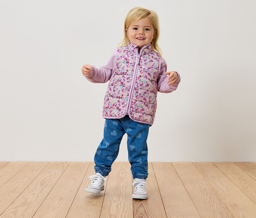 Egy kis szőke lány áll egy fapadlón, virágos mellényt, lila felsőt és farmert visel.