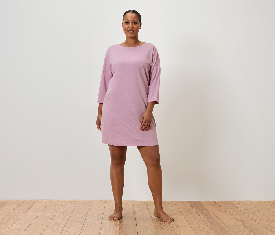 Egy nő áll mezítláb egy fal előtt, és egy rózsaszín, rövid ujjú ruhát visel.