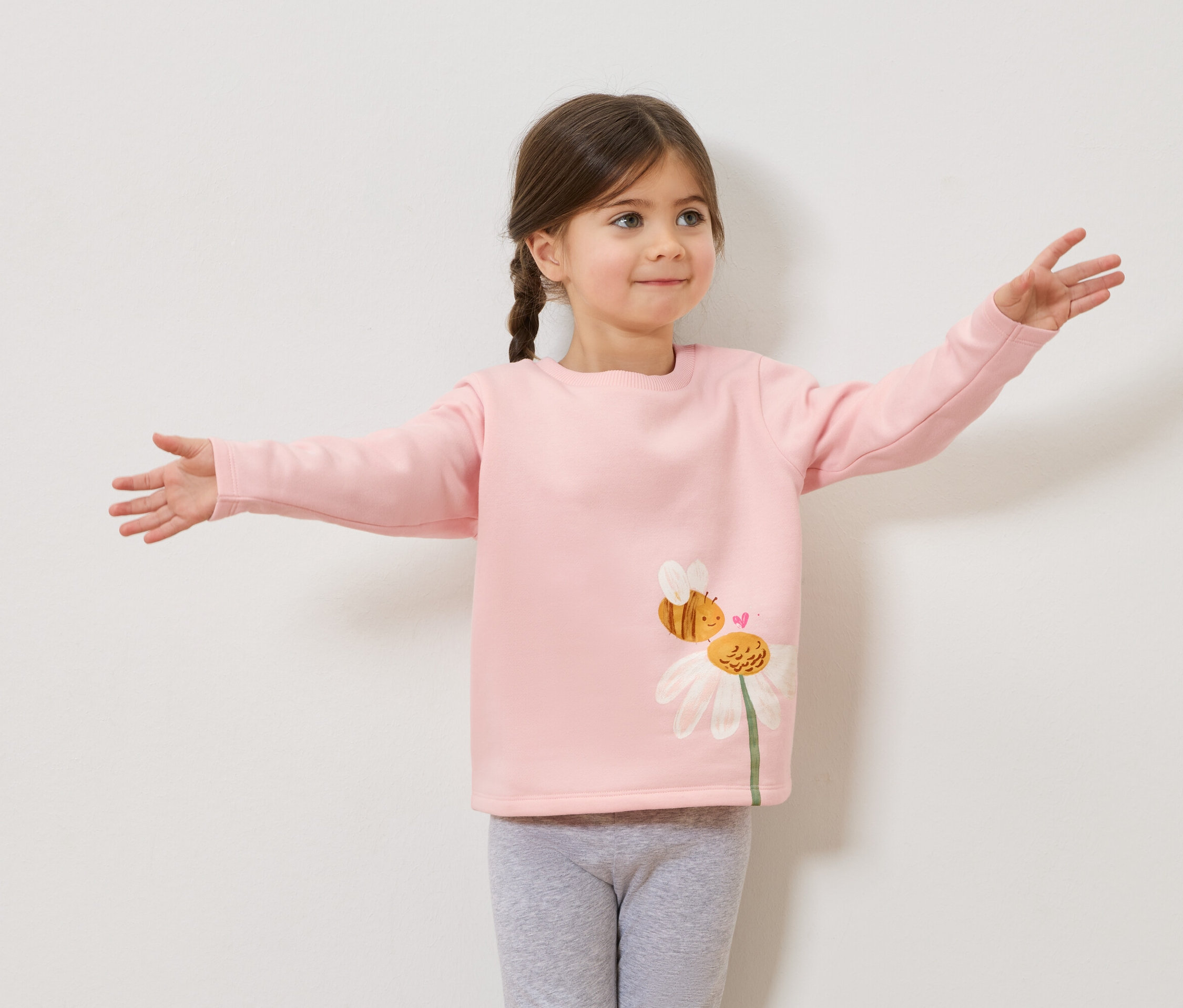 Lány fonott hajjal, rózsaszín, méhecskés és százszorszépes mintás pulóverben, szürke leggingsben, kinyújtott karokkal.