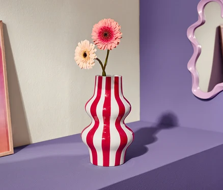 Két rózsaszín gerbera virág egy piros-fehér csíkos vázában egy lila polcon, mellette egy rózsaszín kép és egy ívelt tükör.