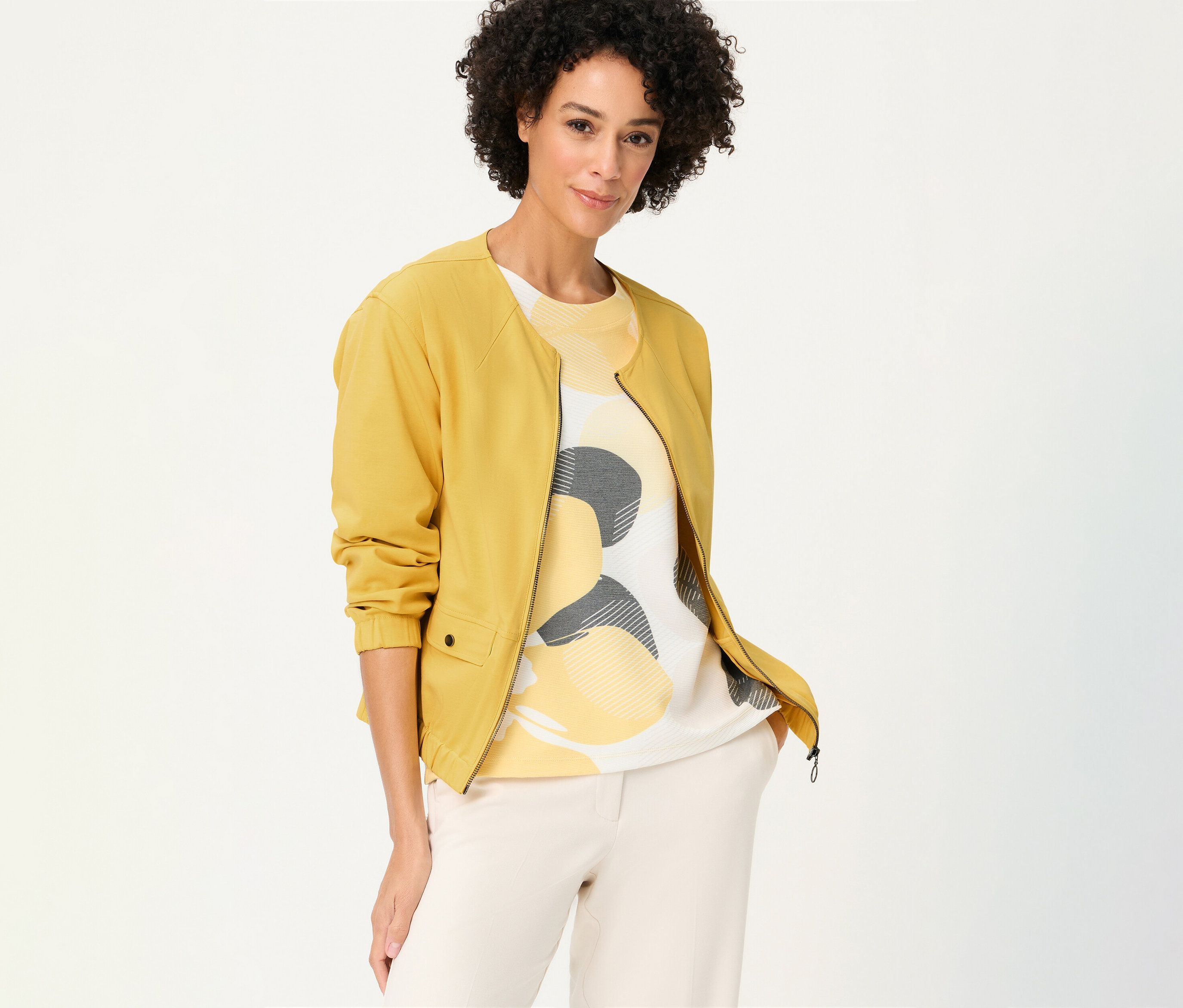 A göndör hajú nő egy mintás felső és világos nadrág fölött egy sárga Olsen szabadidőfelsőt visel.