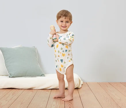 Mosolygó kisfiú levélmintás bodyban csörgőt tart a kezében. A háttérben egy párna és egy futon látható.