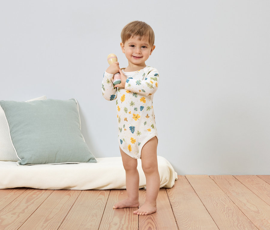 Mosolygó kisfiú levélmintás bodyban csörgőt tart a kezében. A háttérben egy párna és egy futon látható.