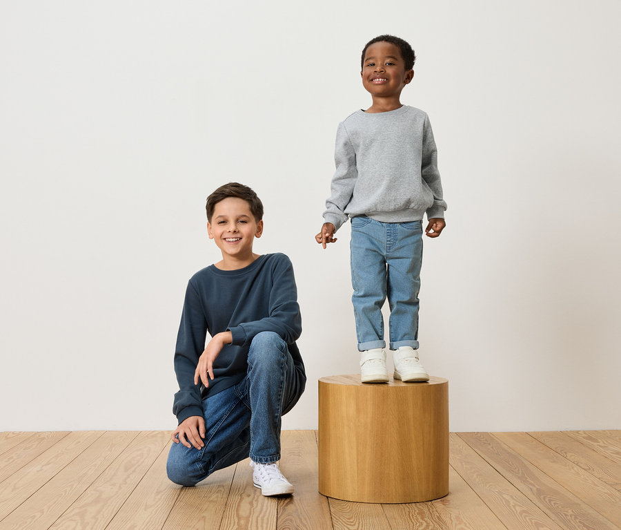 Két fiú pózol: az egyik tengerészkék pulóverben és farmerban guggol, a másik szürke pulóverben és farmerban áll egy fa zsámolyon.