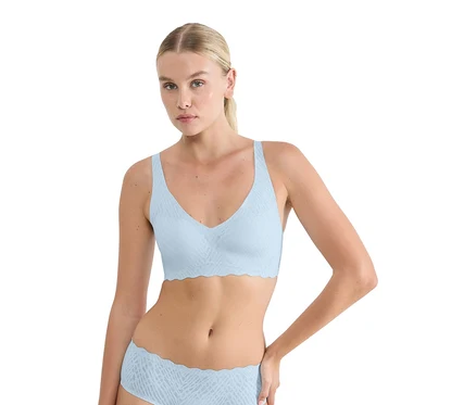 Szőke hajú nő visel egy azúrkék sloggi ZERO Feel Bliss Soft melltartót és hozzá illő alsóneműt finom mintával és hullámos szegéllyel.