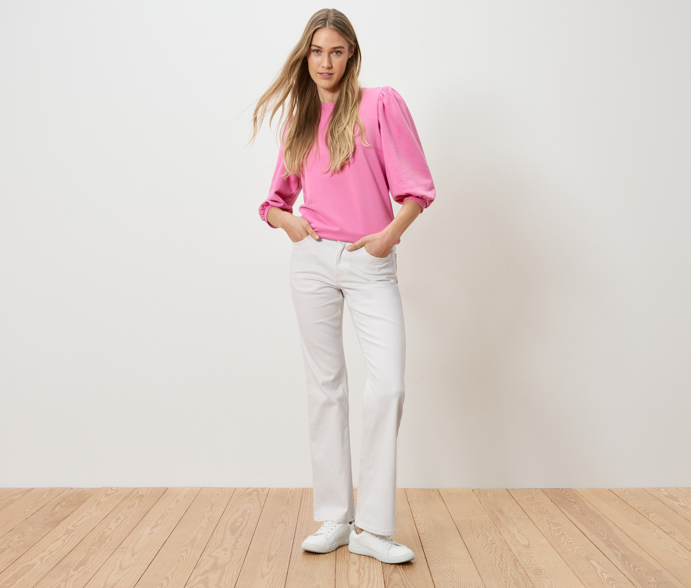 Egy nő áll a kezével a nadrágzsebében. Rózsaszín felsőt és fehér farmert visel.