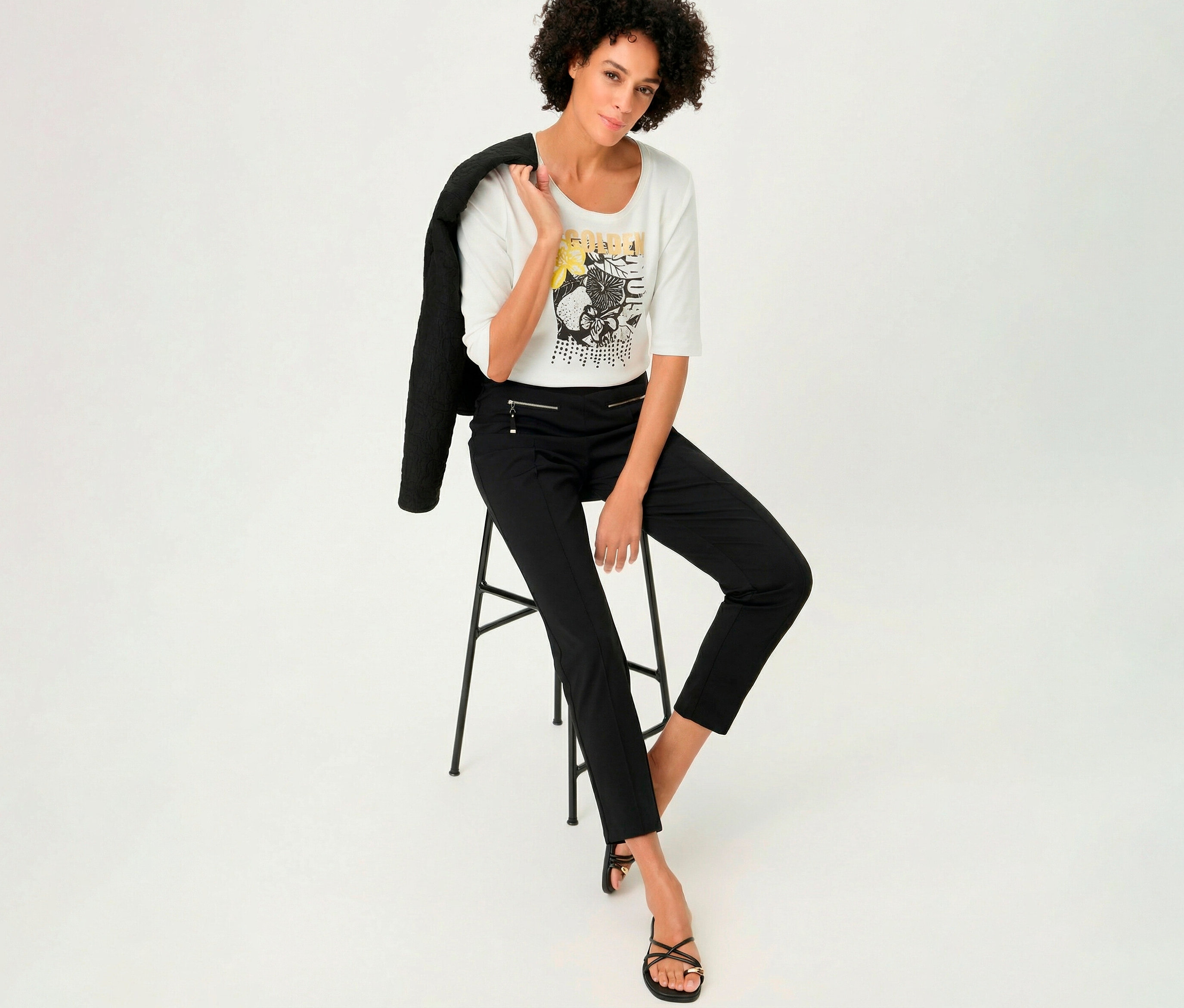 Nő ül egy széken Olsen 3/4-es ujjú pólóban, a vállán zakóval.