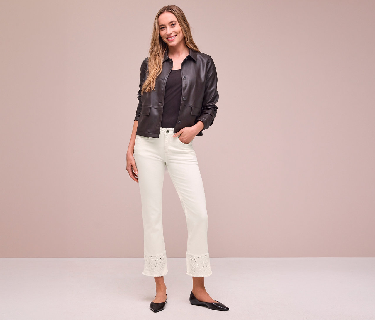 Mosolygó nő sötétbarna dzsekiben, fekete felsőben és világos Street One Jeans-ben perforált szegéllyel, egyik kezével a zsebében áll.