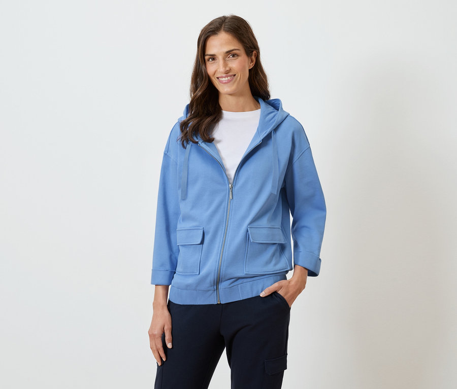 Hosszú barna hajú nő mosolyog kék kapucnis pulóverben és sötét nadrágban.