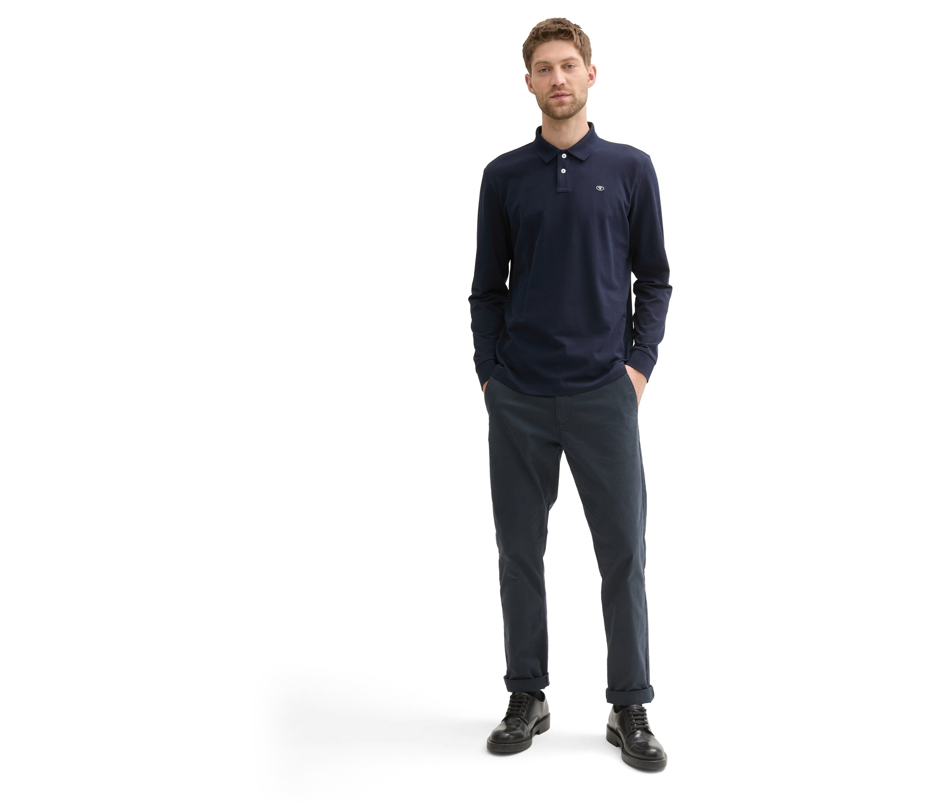 Egy férfi áll, sötétkék, hosszú ujjú pólót, sötétkék nadrágot és fekete cipőt visel.