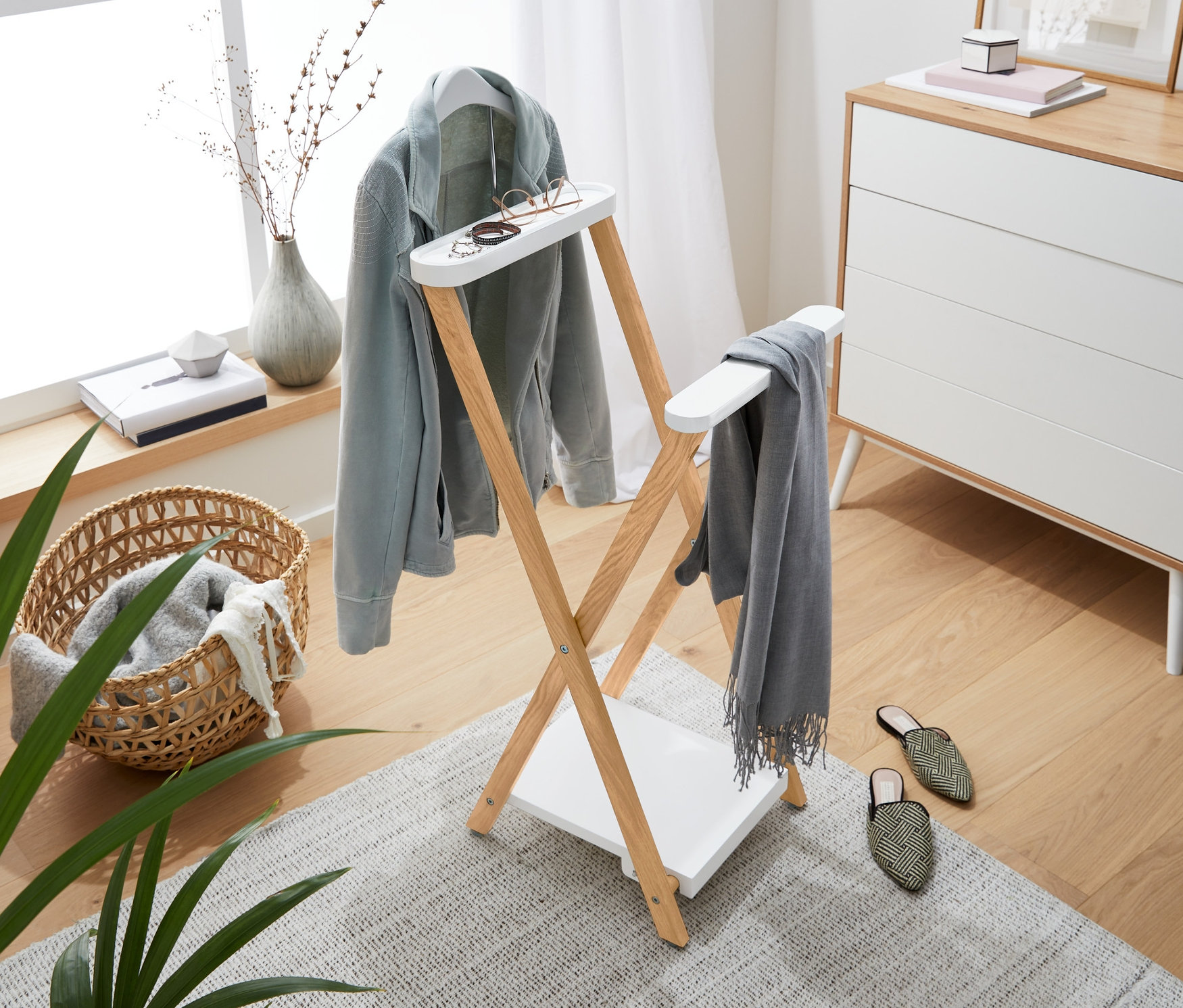 Világos szoba fa ruhaállvánnyal, amin világoskék kabát, szürke sál és szemüveg lóg. A háttérben komód, ablak és dekoráció.