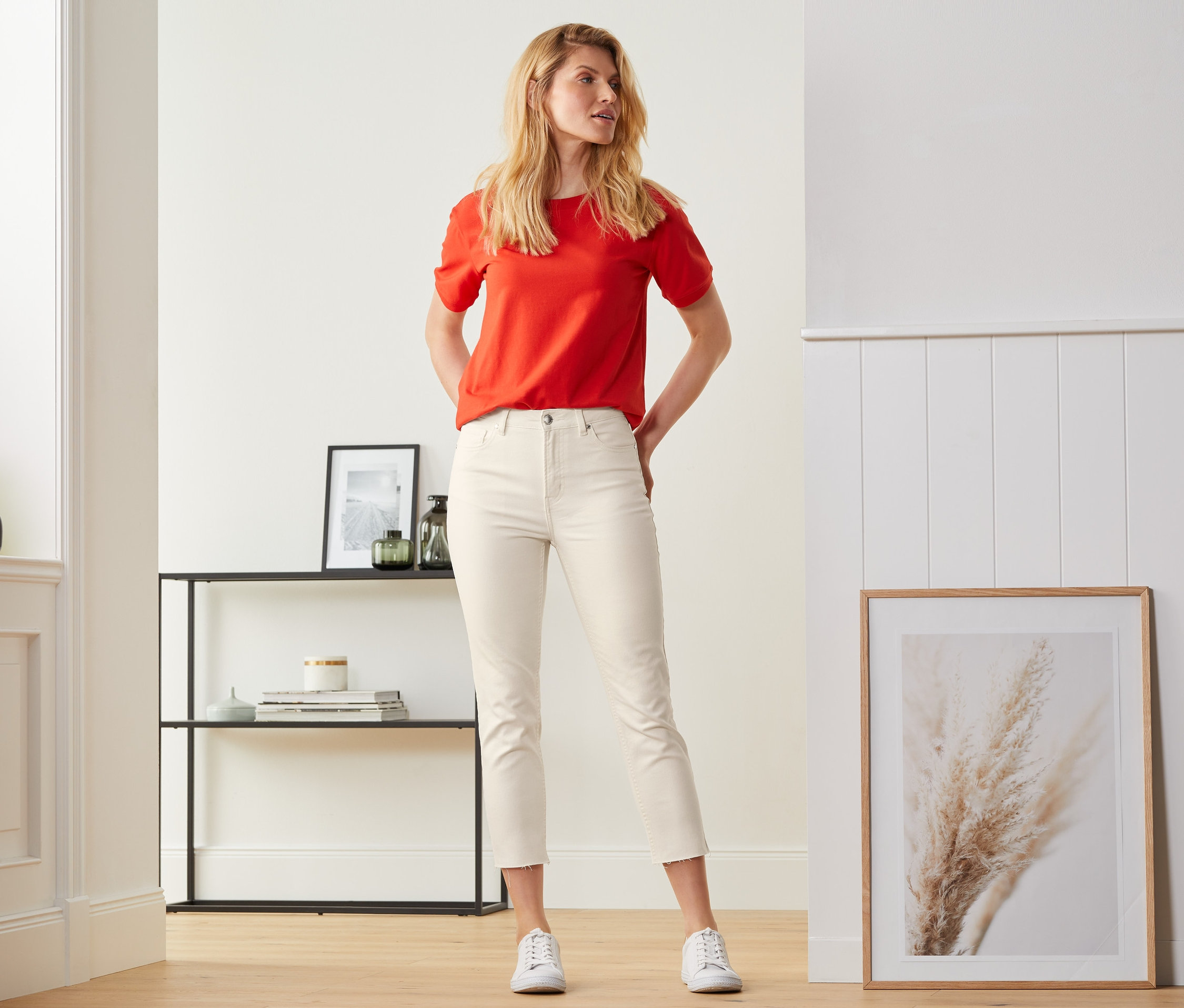 Egy nő pózol törtfehér, 7/8-os farmernadrágban és egy piros felsőben.