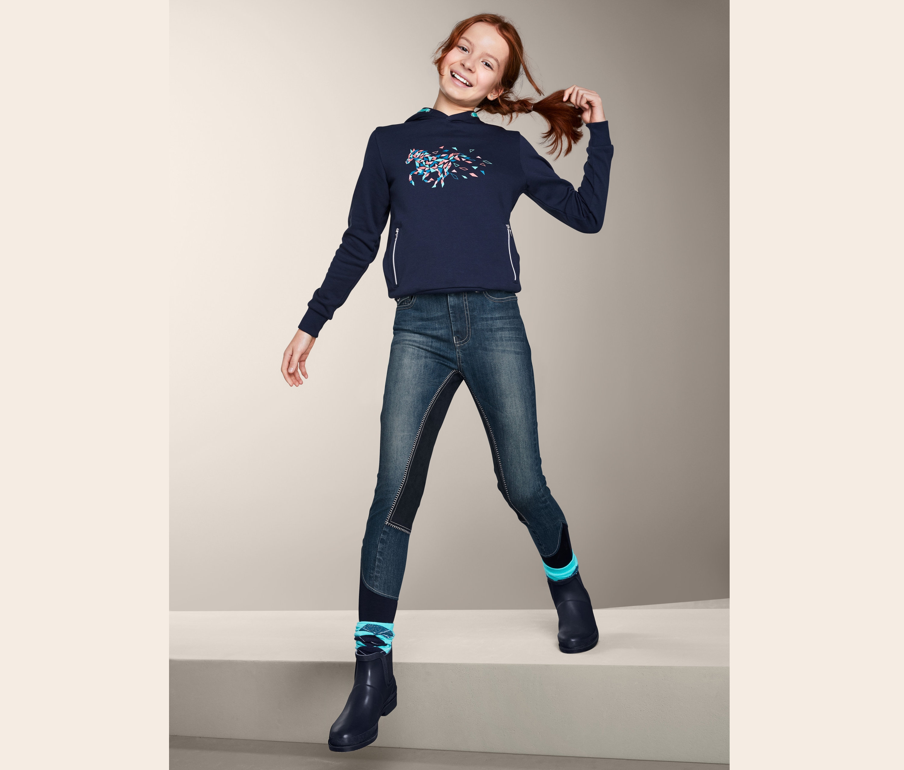 Vörös hajú lány pózol USG lány farmer lovaglónadrágban, kék kapucnis pulóverben és kék gumicsizmában.