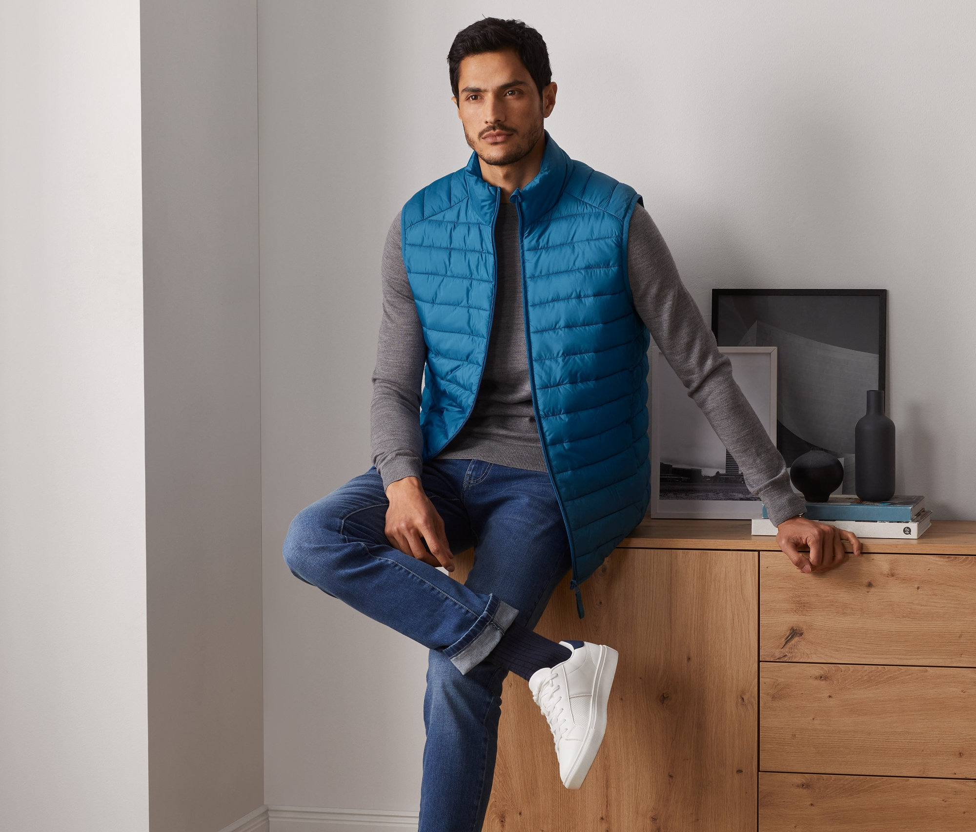 Férfi ül a komódon és kék steppelt mellényt visel.