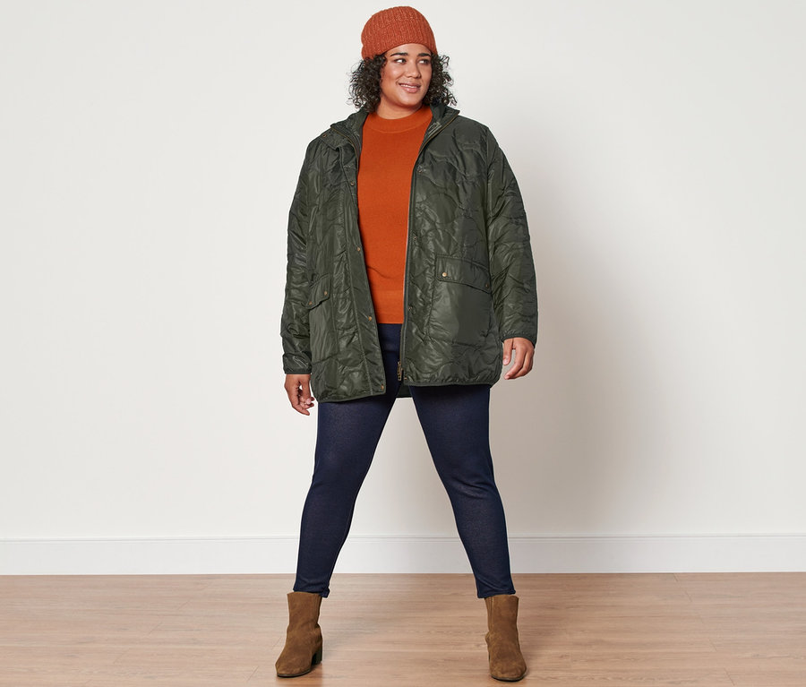 Egy nő kötött pulóvert, steppelt kabátot, farmerhatású bélelt leggingst, kötött sapkát és velúrbőr csizmát viselve pózol a kamerának.