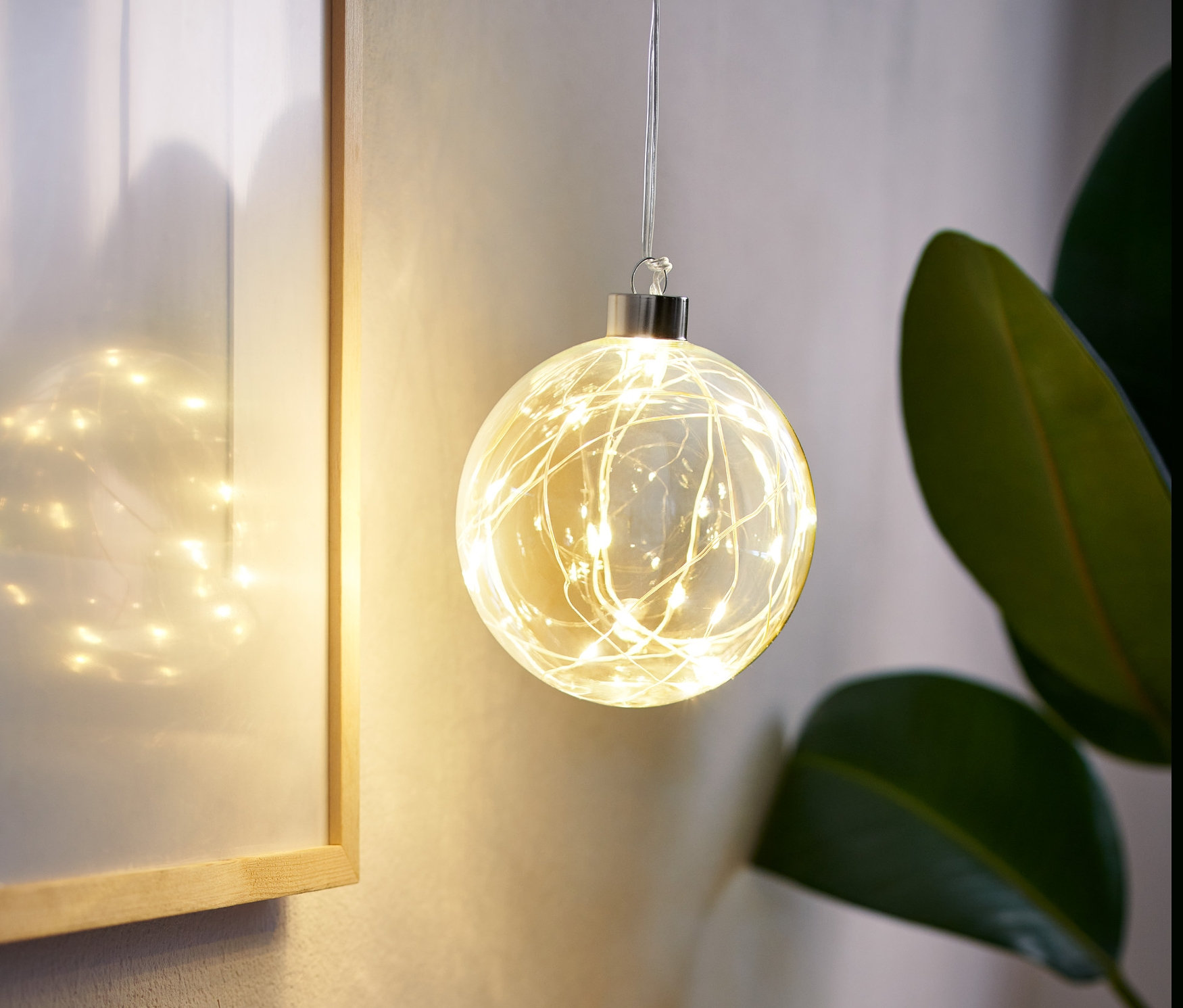 Egy LED-es üveggömb lóg a szobában, mellette egy keretes kép és egy váza fűvel.