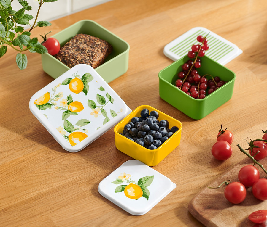 Felülnézet egy fa asztalra, amelyen különböző színű uzsonnás dobozok találhatók gyümölcsökkel és zöldségekkel.