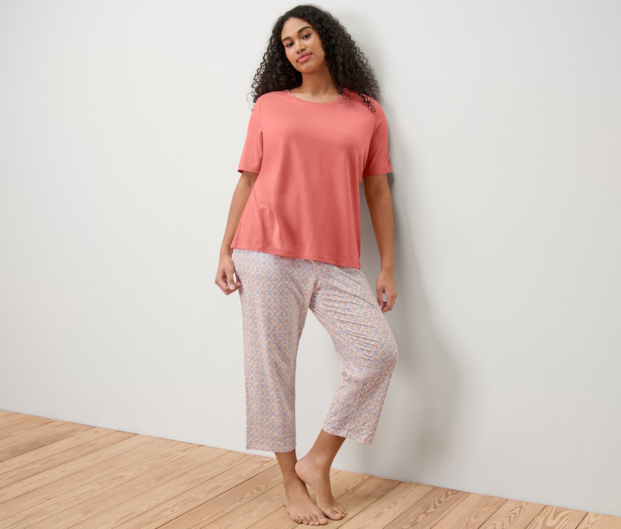 A falnak támaszkodó nő lazacszínű pizsama pólót és mintás 7/8-os pizsamanadrágot visel.