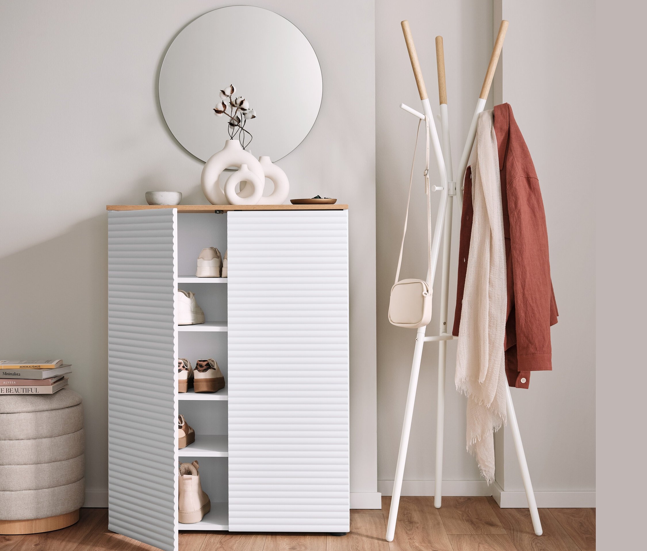 Cipősszekrény nyitott ajtóval, ruhafogas és kerek puff tárolóval mellett.