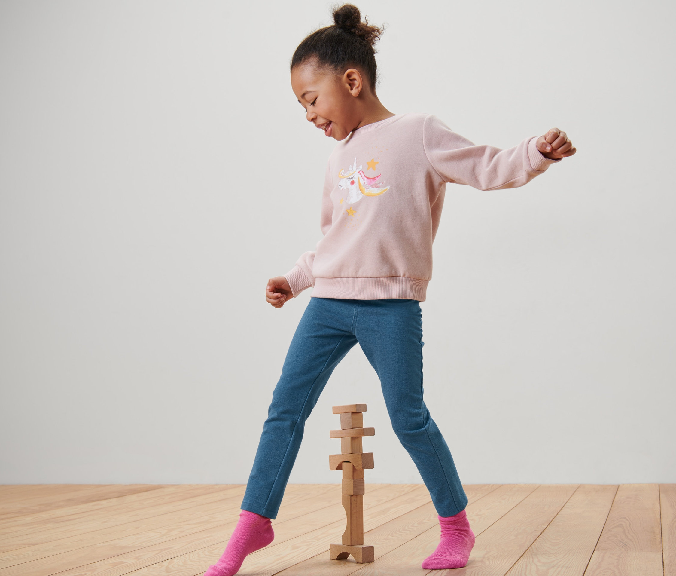 Egy kislány rózsaszín unikornisos pulóverben és kék »Mia« jeggingsben játszik fakockákkal.