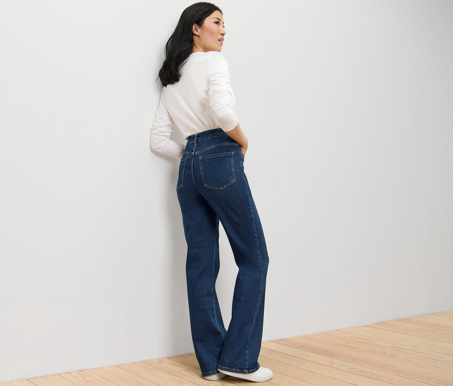 Egy nő sötétkék, Lou szabású bootcut farmerban és fehér, hosszú ujjú felsőben pózol.