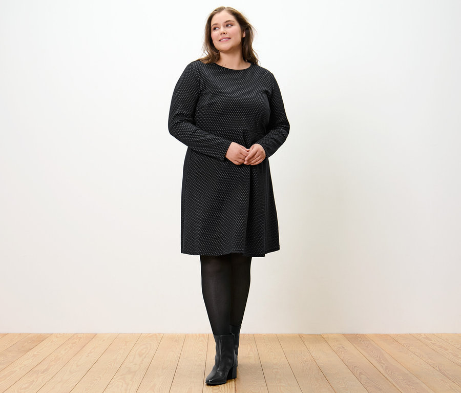 Nő áll, csillogó jacquard ruhát visel.