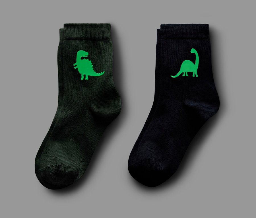 Két pár zokni, egy sötétzöld dinoszaurusz mintával, egy fekete dinoszaurusz mintával, mindkét pár 'Glow in the Dark'.