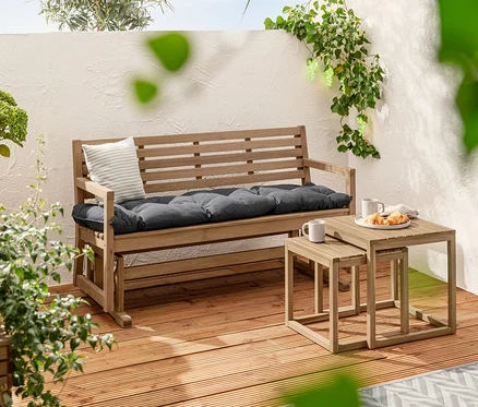 A »Lenja« kisasztal-szett egy Sun Garden kertipad-párna, kb. 160 x 50 cm méretű pad mellett áll. A háttérben 2 prémium napelemes leszúrható lámpa és egy szőlő alakú napelemes fényfüzér látható.