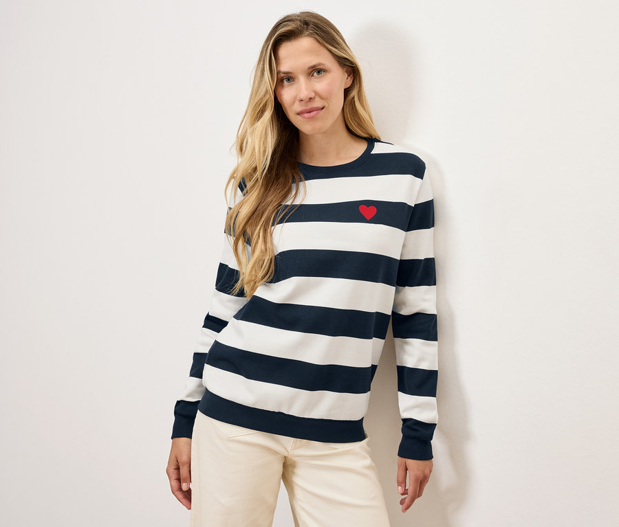 Szőke nő pózol egy piros szívvel díszített, csíkos pulóverben és világos nadrágban.