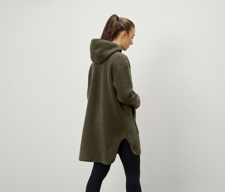 Hátulnézetből egy nő olívazöld, bolyhos kapucnis kabátot és fekete leggingset visel.