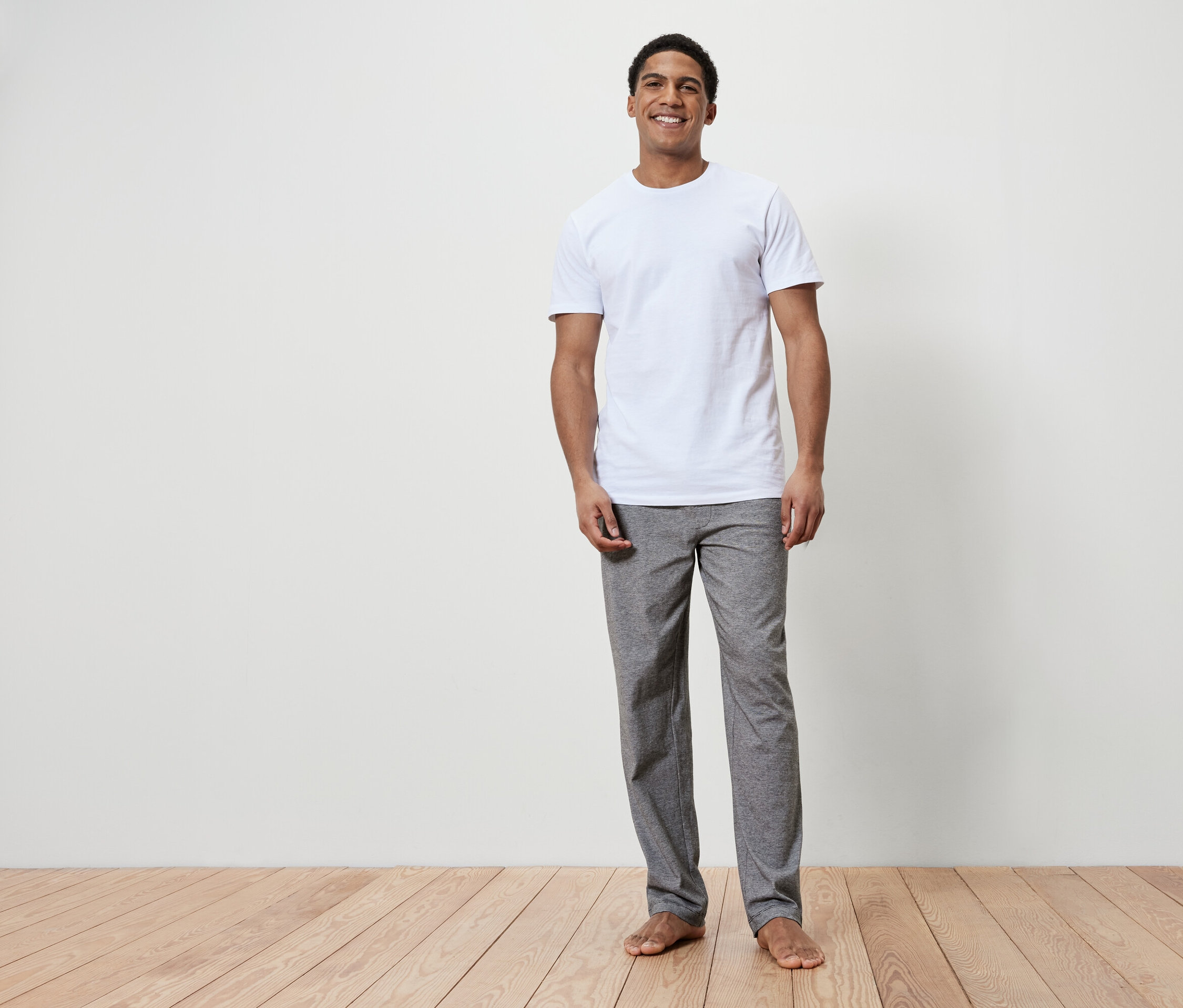 Egy mosolygó férfi áll mezítláb egy fapadlón. Fehér pólót és szürke nadrágot visel.