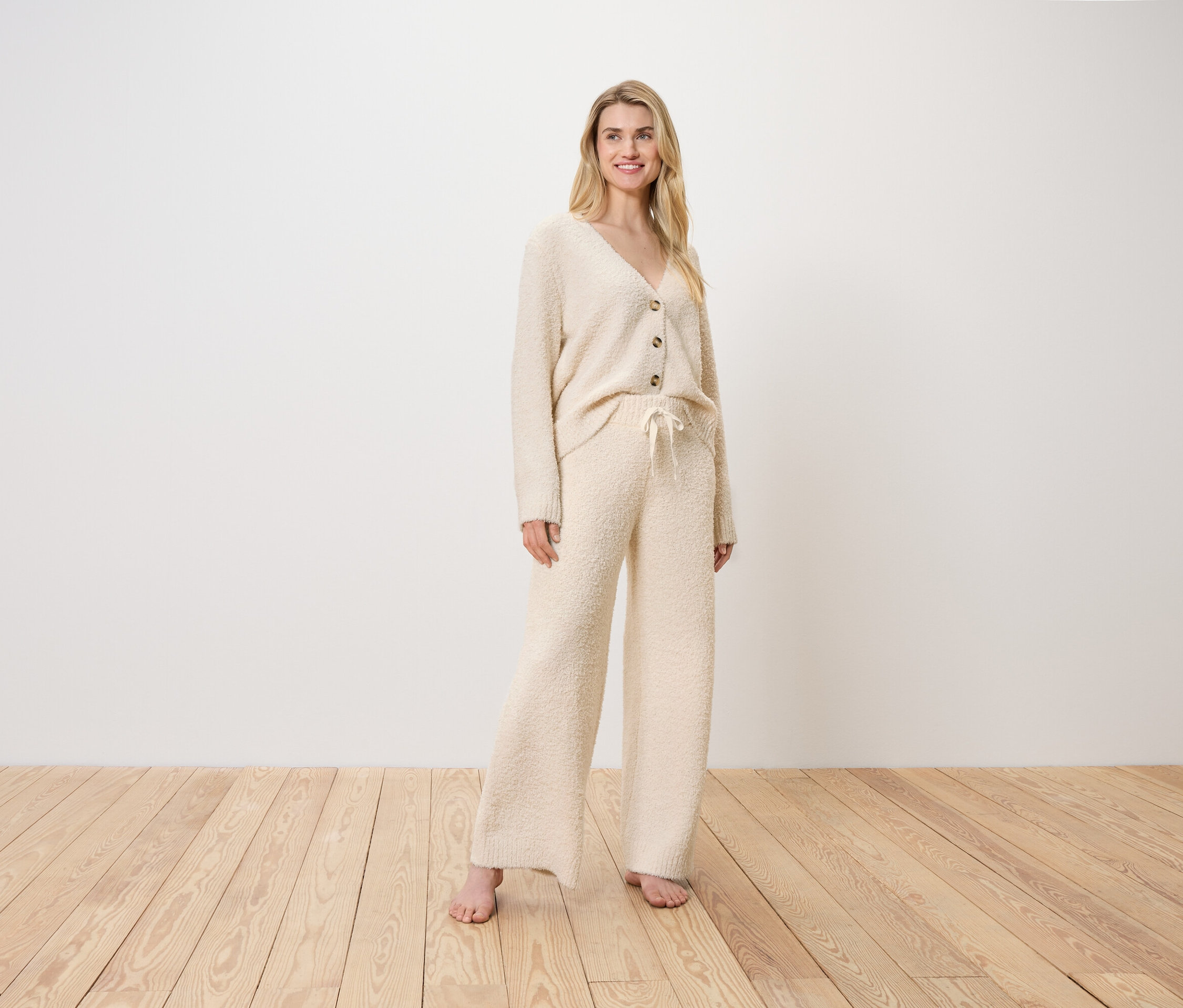 Egy szőke nő krém színű kardigánban és nadrágban áll a fapadlón.