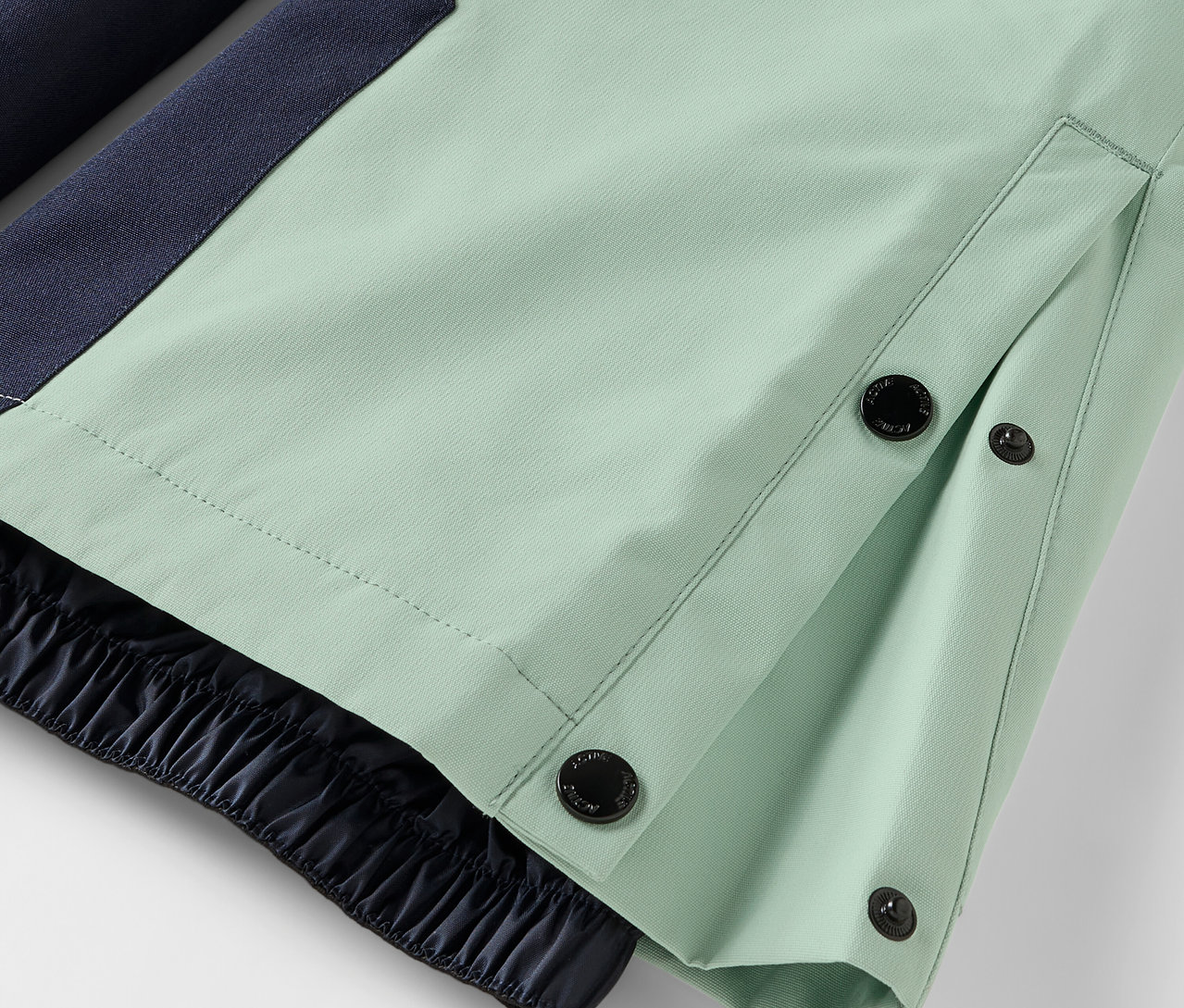 Világoszöld nadrág részlete gombos zárással és rugalmas szegéllyel.