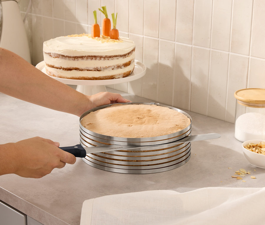 Egy személy egy tortát vág egy állítható tortaszeletelővel. A háttérben egy torta sárgarépával és üvegek fedővel.