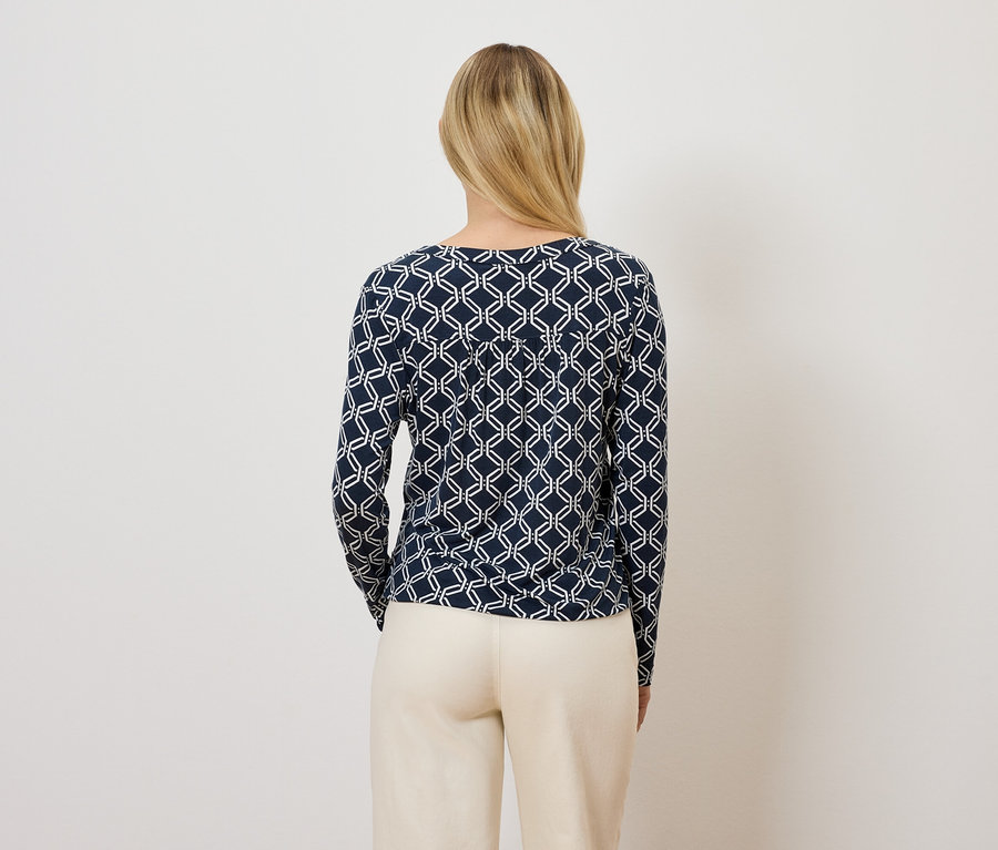 A fényképezőgépnek háttal álló nő sötétkék, geometrikus mintás, hosszú ujjú felsőt és bézs nadrágot visel.