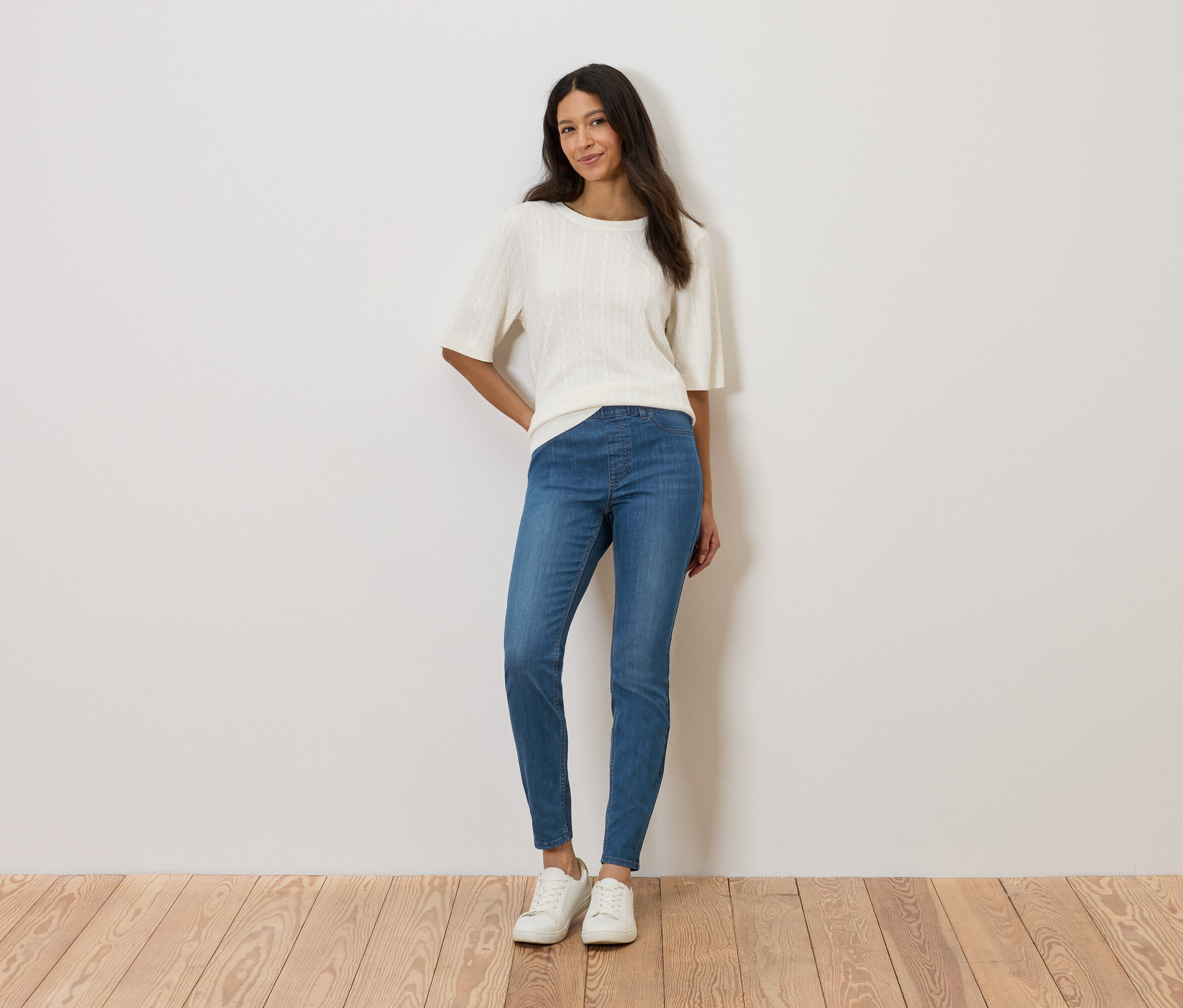 Nő áll a fal előtt, fehér felsőt, kék farmert és fehér cipőt visel.