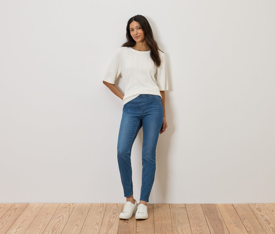 Nő áll a fal előtt, fehér felsőt, kék farmert és fehér cipőt visel.