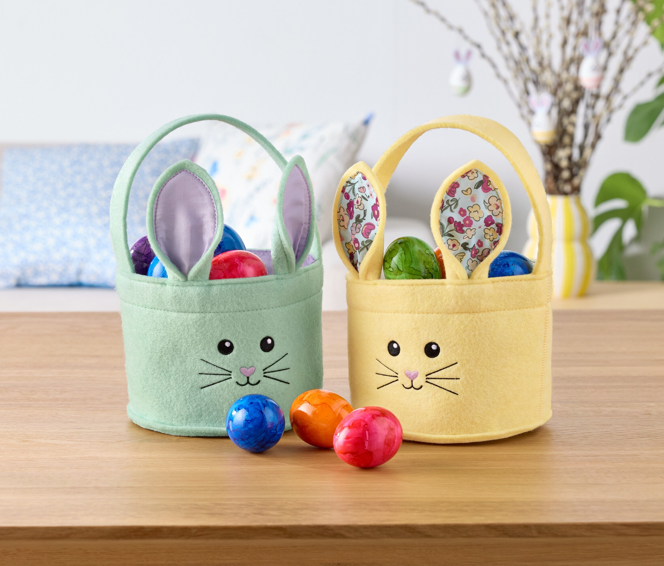 Két húsvéti nyuszi kosár, egy mentazöld és egy sárga, tele színes tojásokkal, áll egy asztalon.
