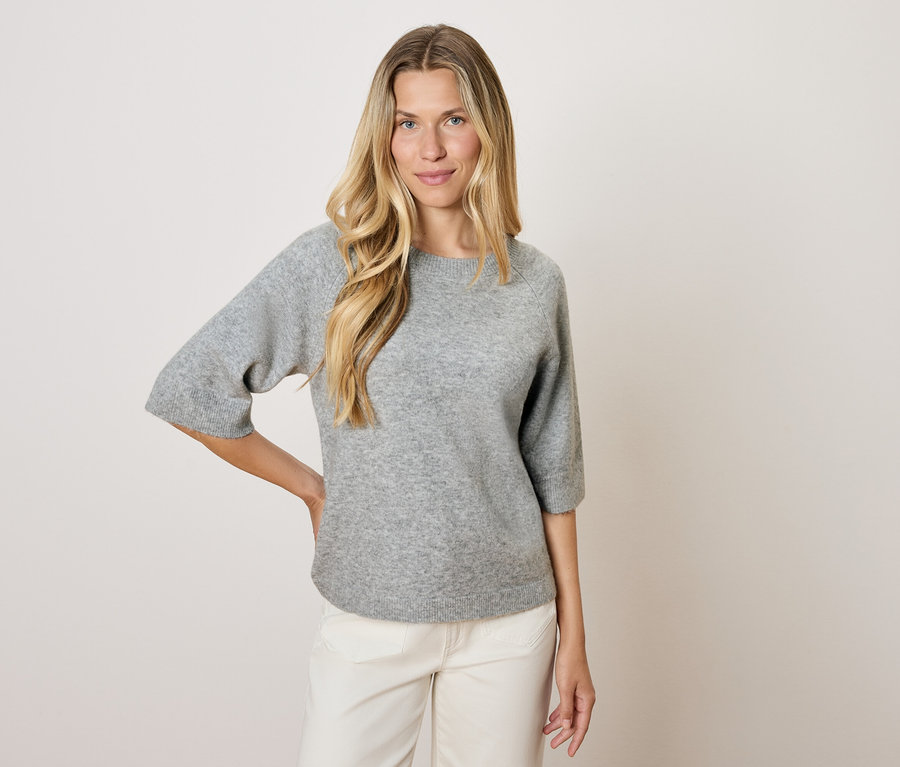Egy szőke nő háromnegyedes ujjú, szürke pulóverben és fehér nadrágban pózol világos háttér előtt.