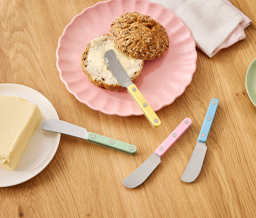 Vajas zsemle rózsaszín tányéron egy vajtömb és színes kések mellett egy fa asztalon.