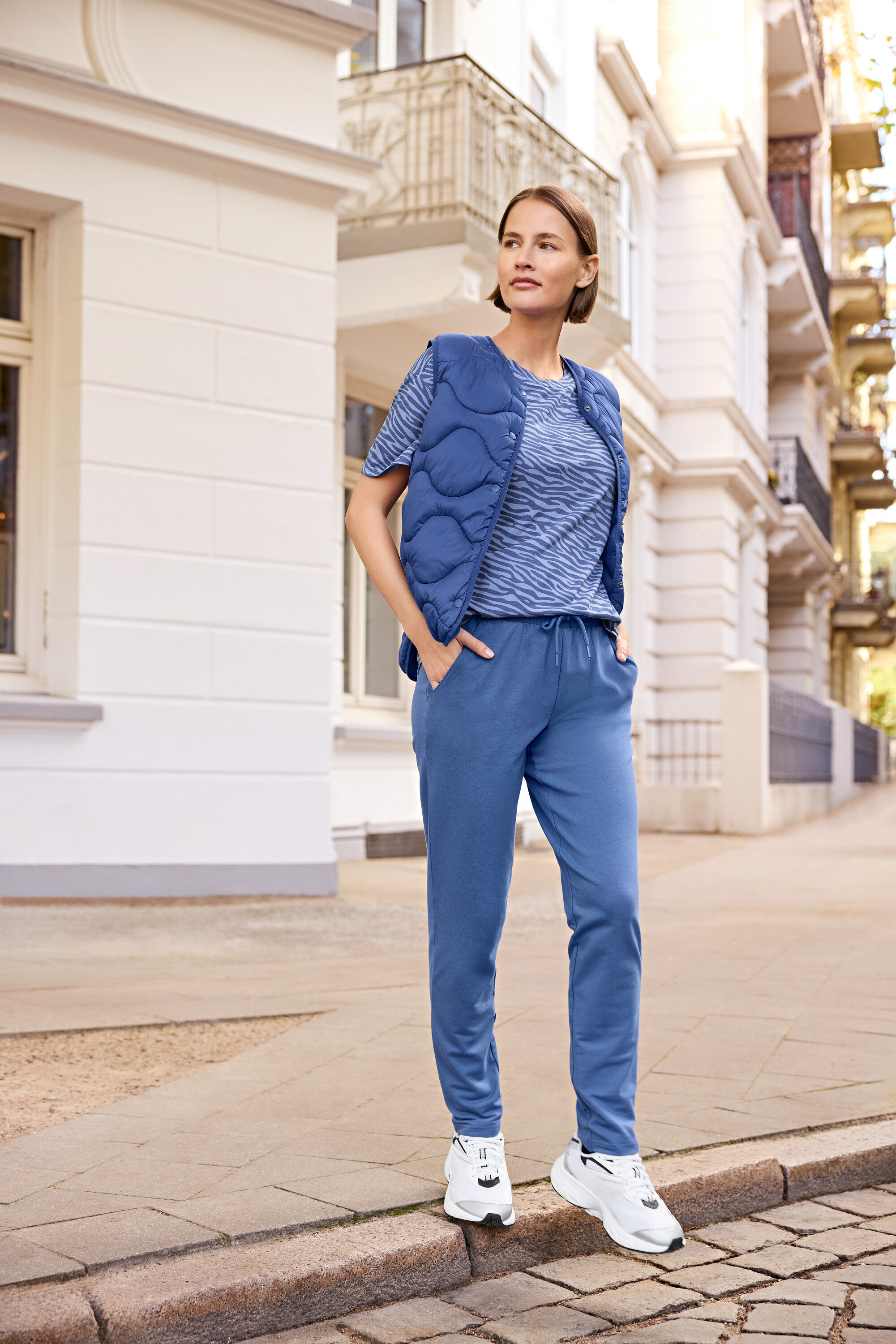 Nő áll a járdán, kék mellényt, kék zebra mintás pólót és kék melegítőnadrágot visel.