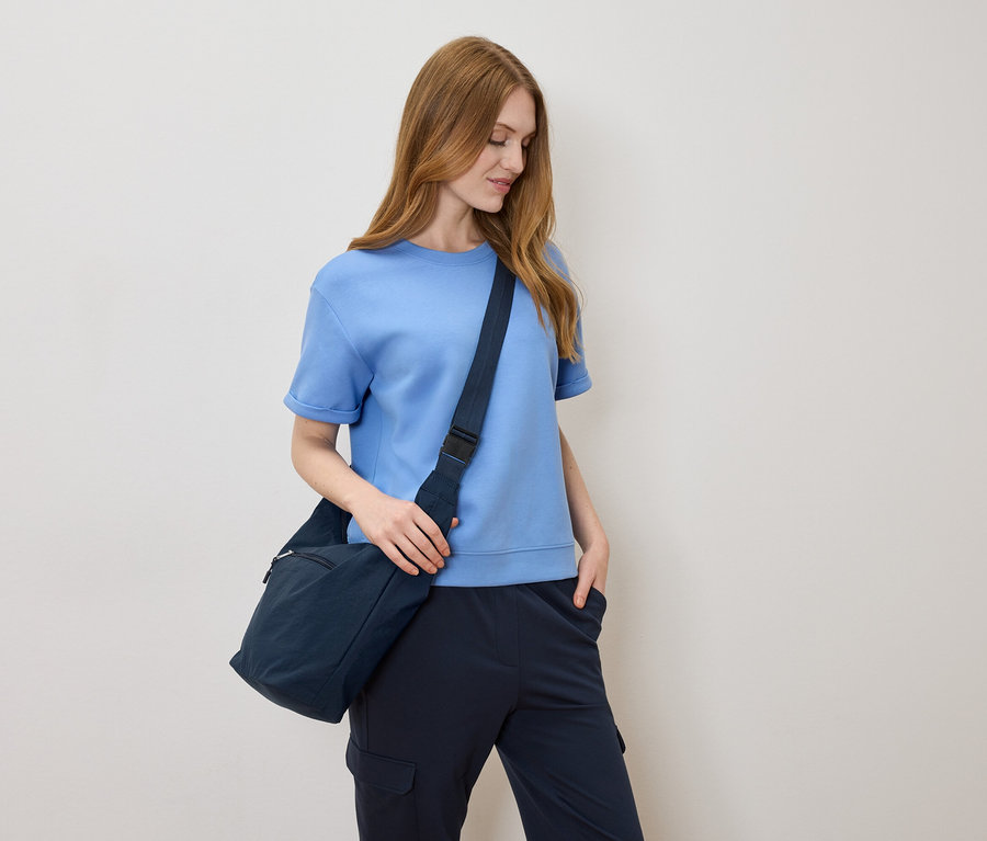 Világosbarna hajú nő világoskék pólóban és tengerészkék válltáskával, krémszínű háttér előtt.